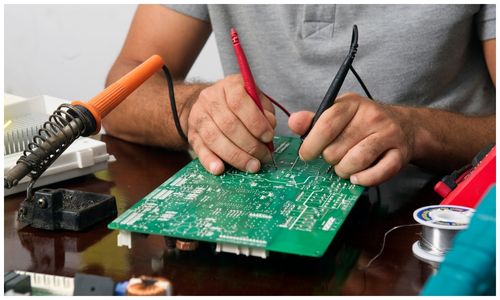 Meter probes held against a decoder circuit board on a repair bench, soldering iron alongside